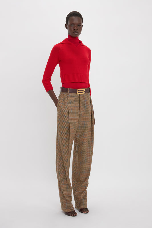 A person stands against a plain background wearing a Double Layer Top In Deep Red by Victoria Beckham, brown belt, and high-waisted plaid trousers.