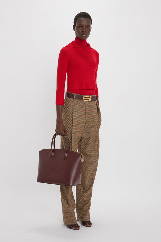 Person wearing a red Victoria Beckham Double Layer Top In Deep Red and brown checkered pants, holding a burgundy handbag and standing against a white background.