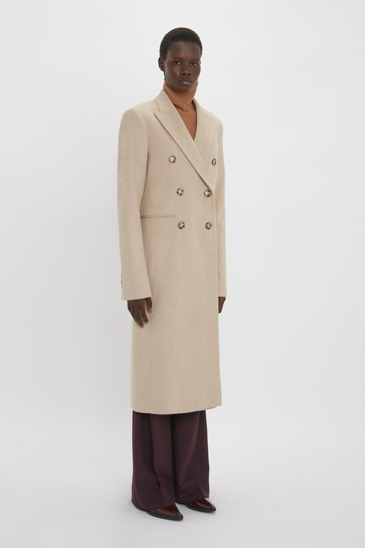 A person stands wearing a long, Tailored Slim Coat In Bone by Victoria Beckham over a brown shirt and dark trousers. They have short hair and are posing against a plain white background.