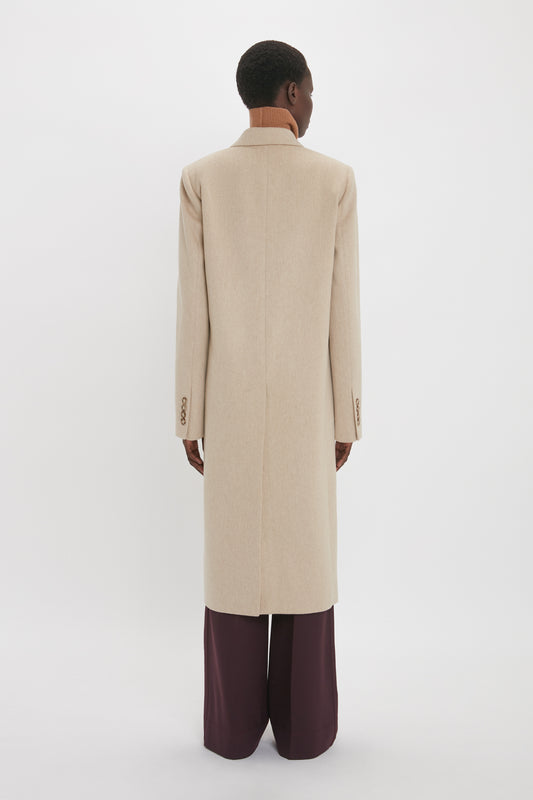 Rear view of a person wearing the Victoria Beckham Tailored Slim Coat In Bone over a brown turtleneck and dark wide-leg pants, standing against a plain white background.