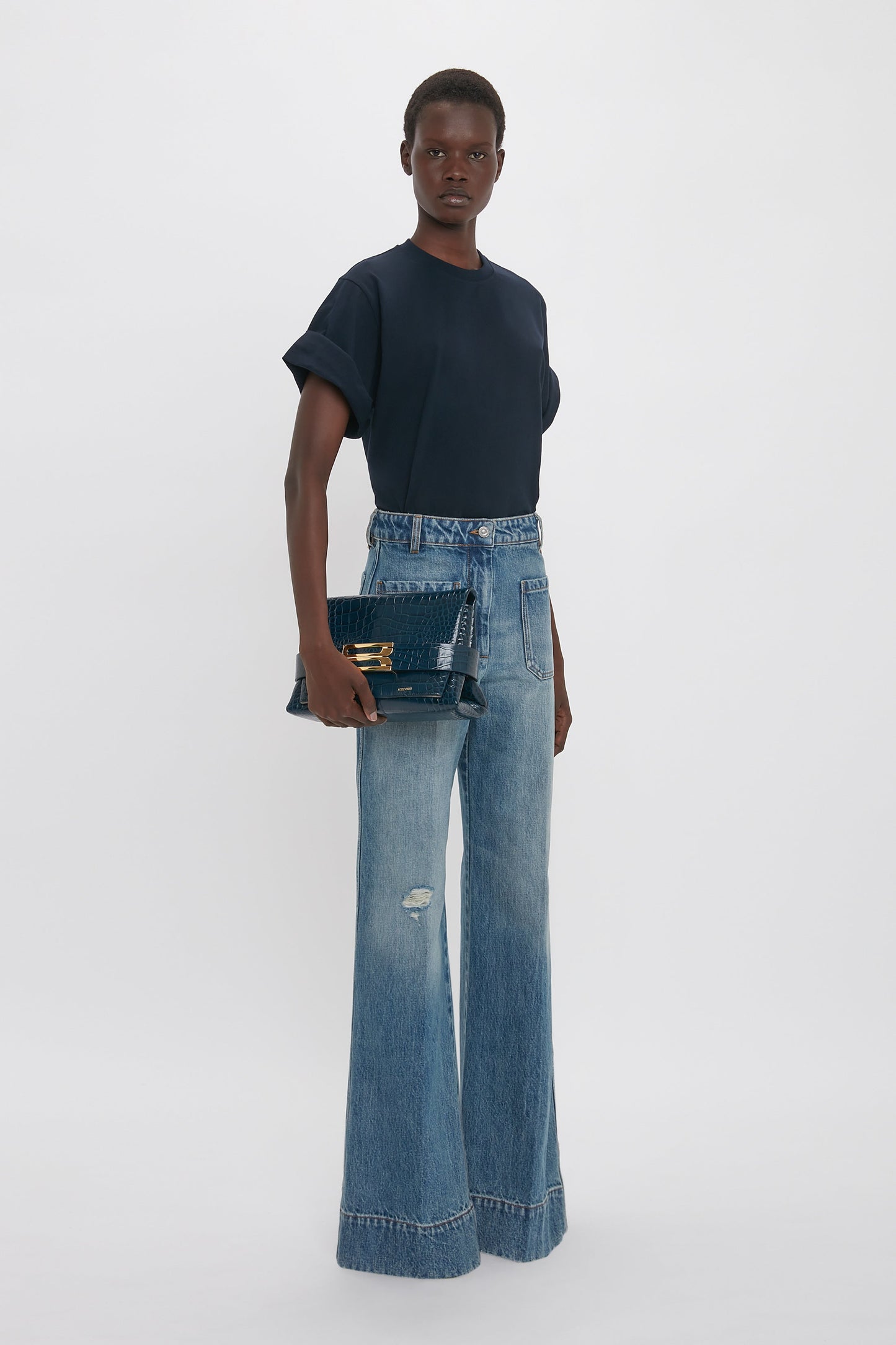 A person in a navy t-shirt and wide-leg distressed jeans holds a B Pouch Bag In Croc Effect Midnight Blue Leather by Victoria Beckham, standing against a plain white background.