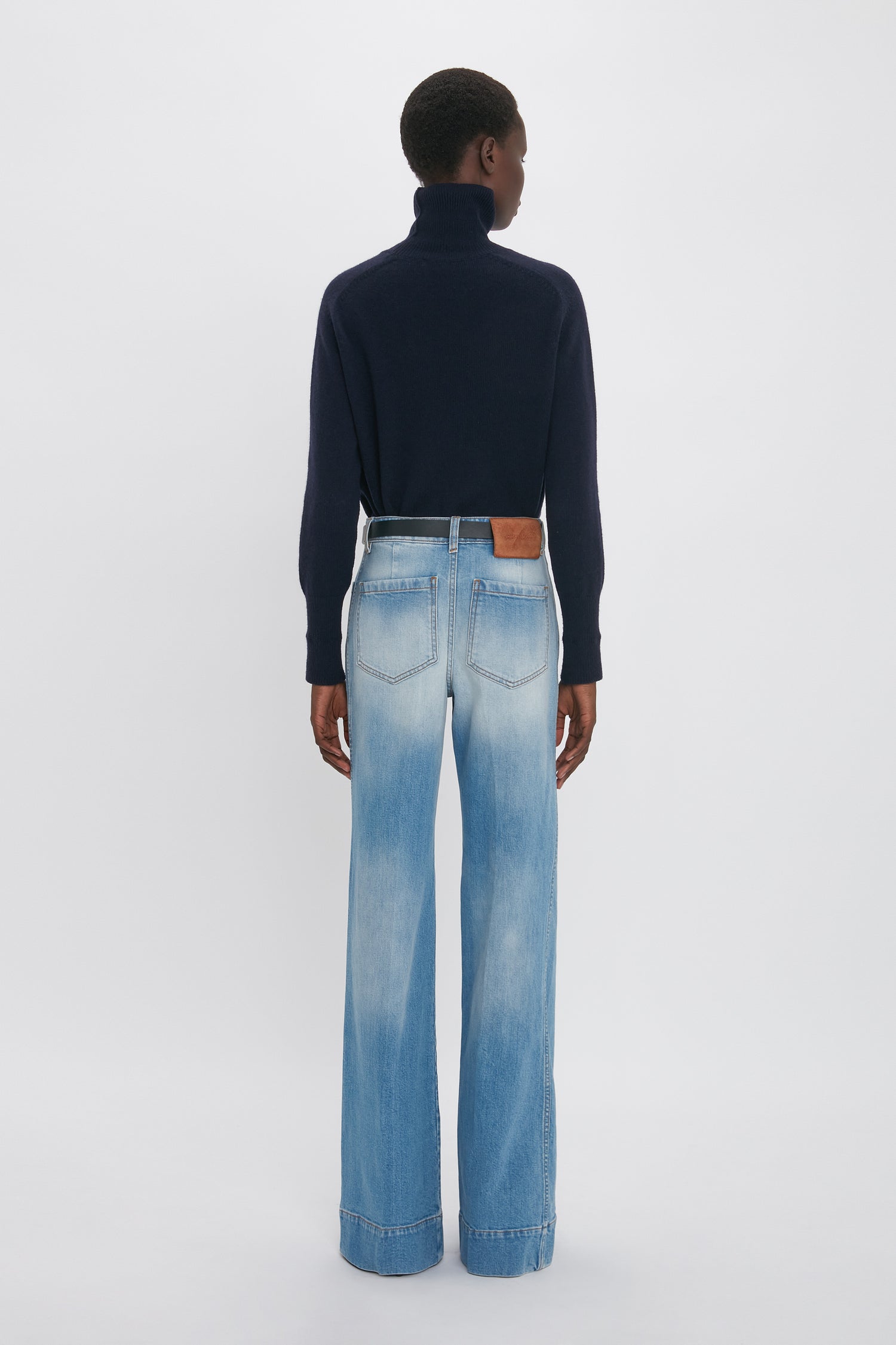 Rear view of a person with short hair wearing a navy turtleneck sweater and stylish tailored Alina High Waisted Jean In Light Summer Wash by Victoria Beckham against a plain white background, exuding an understated elegance reminiscent of Victoria Beckham's fashion.