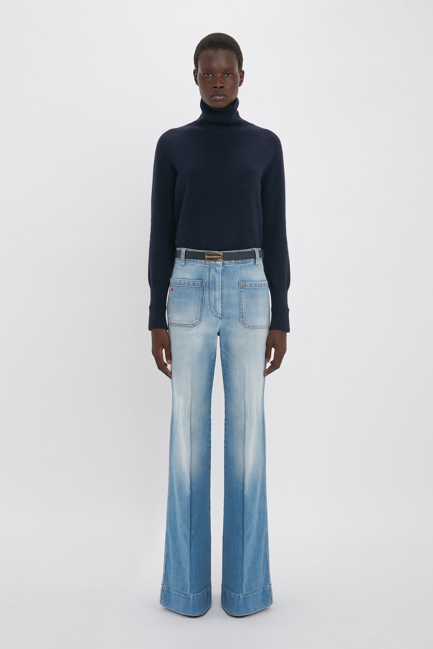 A person stands against a plain background wearing a dark turtleneck sweater and Victoria Beckham Alina High Waisted Jean In Light Summer Wash.