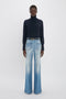 A person stands against a plain background wearing a dark turtleneck sweater and Victoria Beckham Alina High Waisted Jean In Light Summer Wash.