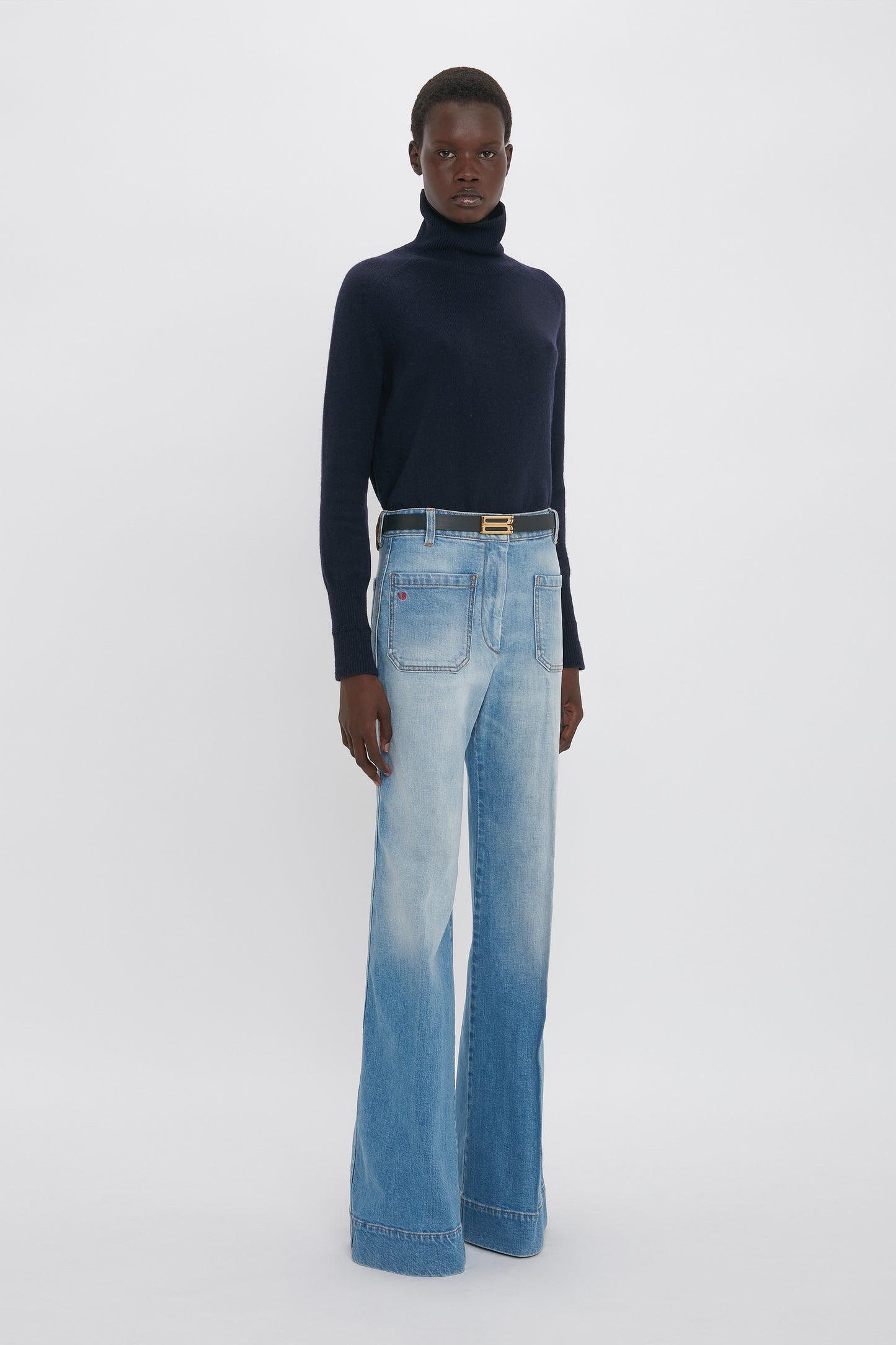 A person stands against a plain background wearing a navy turtleneck sweater, Alina High Waisted Jean In Light Summer Wash by Victoria Beckham, and a black belt.