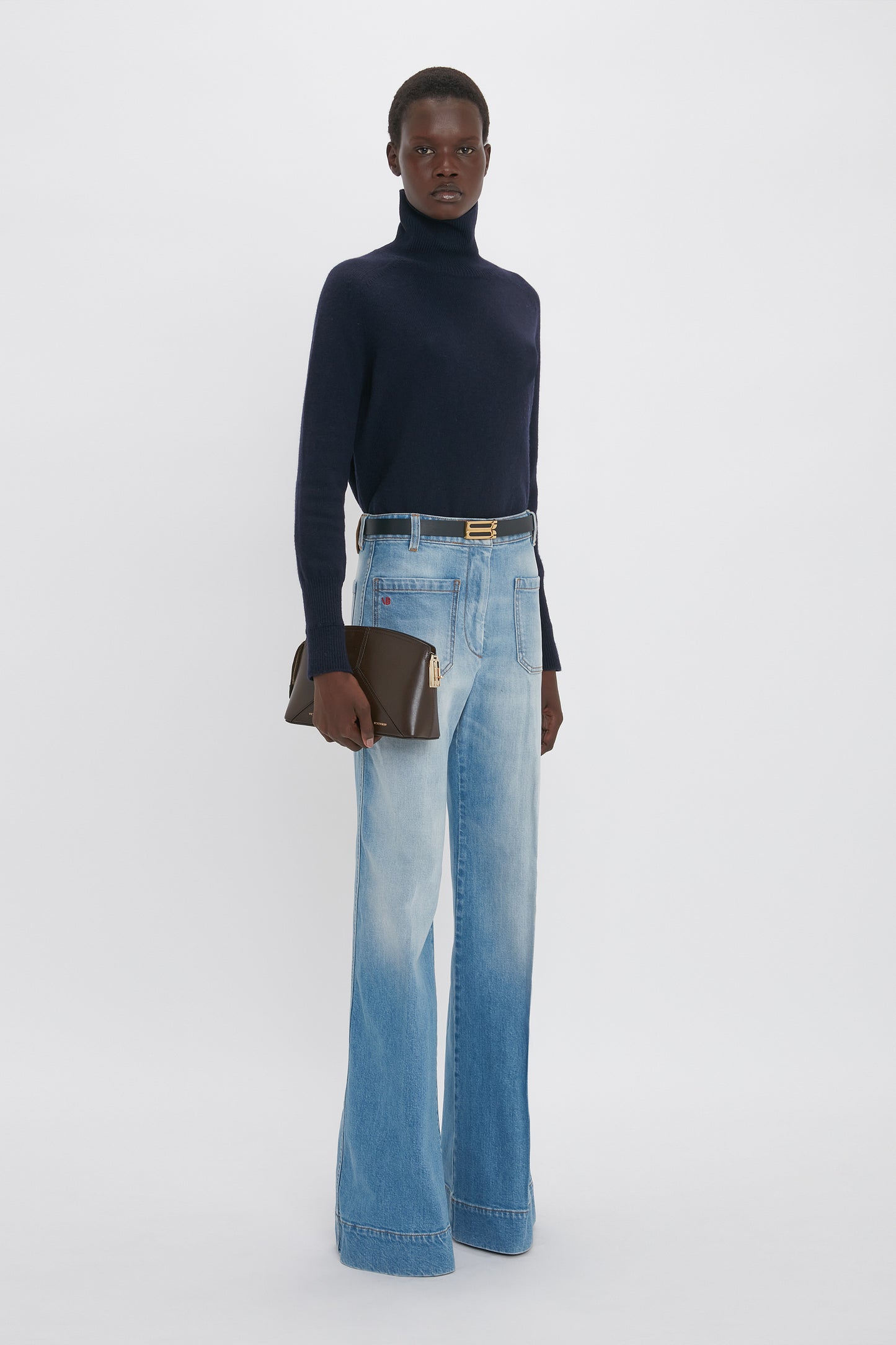 A person stands against a plain background, wearing a black turtleneck, Alina High Waisted Jean In Light Summer Wash from Victoria Beckham that brings to mind Victoria Beckham's sophisticated style, and holding a brown clutch.