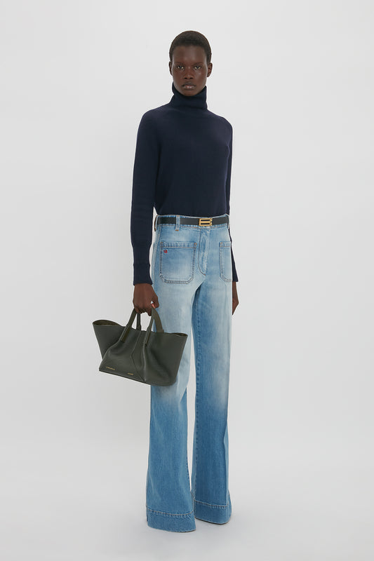 Person standing against a plain white background, wearing a navy turtleneck, light blue flared jeans, and holding a dark green W11 Mini Tote Bag In Loden Leather by Victoria Beckham.
