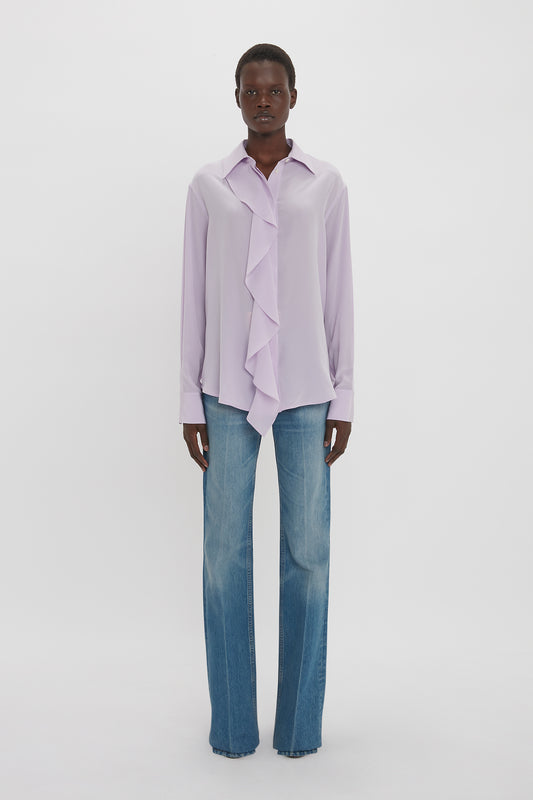 A person stands against a plain white background wearing an Asymmetric Ruffle Blouse In Petunia by Victoria Beckham, paired with blue jeans.