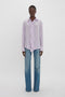 A person stands against a plain white background wearing an Asymmetric Ruffle Blouse In Petunia by Victoria Beckham, paired with blue jeans.