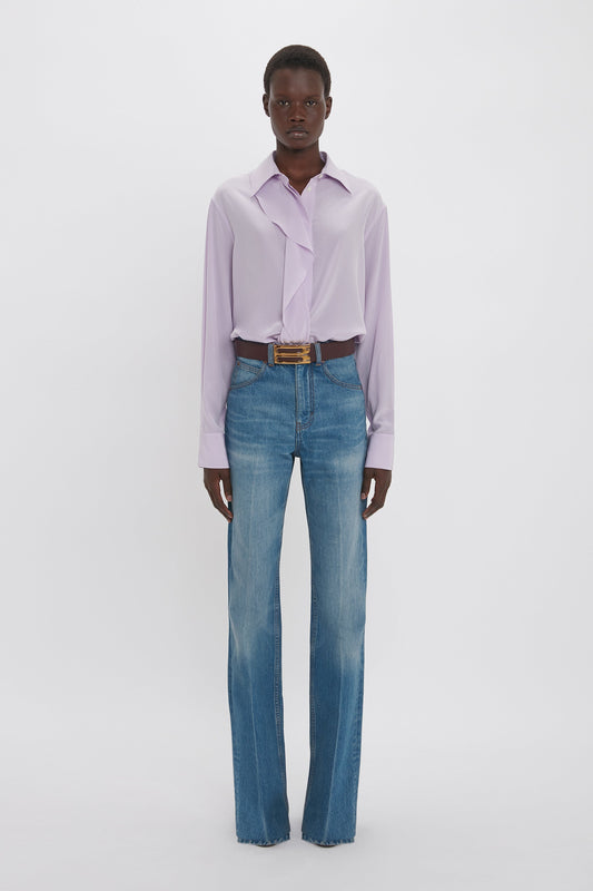 A person wearing a Victoria Beckham Asymmetric Ruffle Blouse In Petunia with blue high-waisted jeans stands against a plain white background, showcasing contemporary contrast.