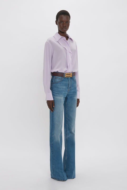 A person stands against a white background, wearing an Asymmetric Ruffle Blouse In Petunia by Victoria Beckham that adds contemporary contrast to their blue jeans and brown belt.