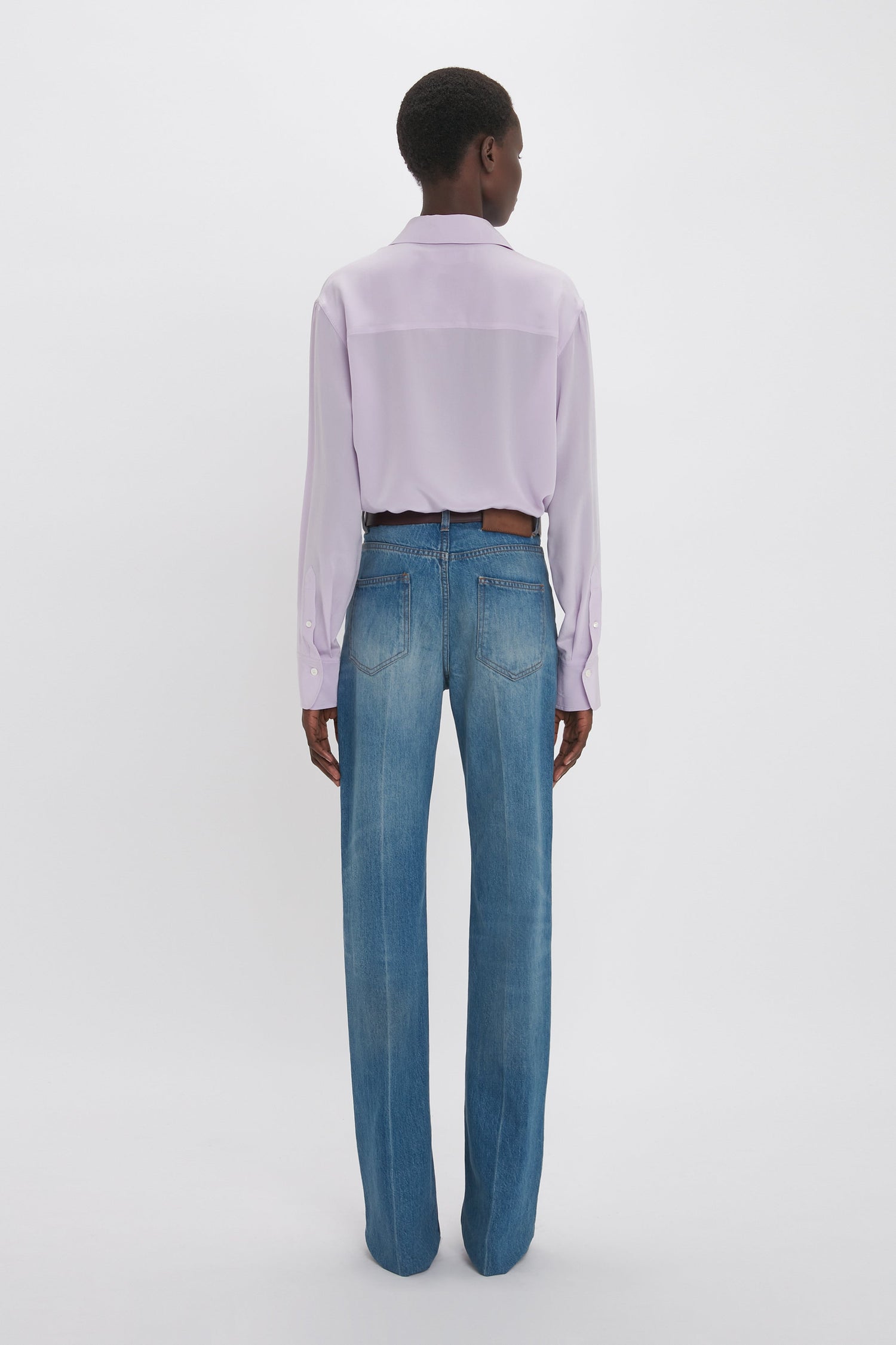 Person standing with their back to the camera, wearing a light purple Asymmetric Ruffle Blouse In Petunia by Victoria Beckham and blue jeans, against a plain white background.