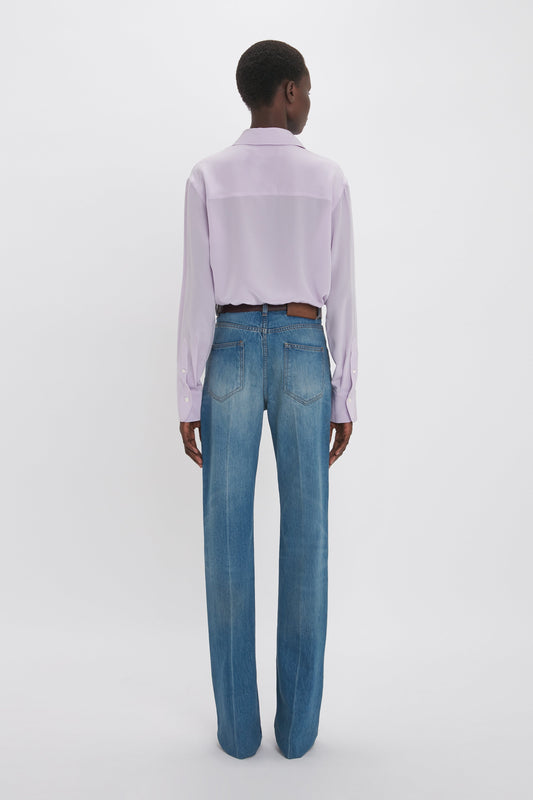 Person standing with their back to the camera, wearing a light purple Asymmetric Ruffle Blouse In Petunia by Victoria Beckham and blue jeans, against a plain white background.