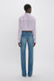 Person standing with their back to the camera, wearing a light purple Asymmetric Ruffle Blouse In Petunia by Victoria Beckham and blue jeans, against a plain white background.