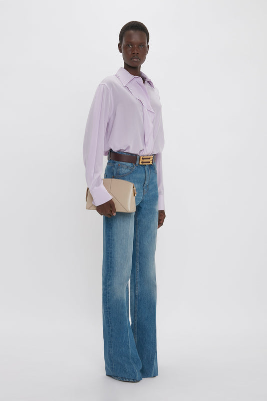A person stands against a plain backdrop, dressed in a light purple Asymmetric Ruffle Blouse In Petunia by Victoria Beckham paired with blue jeans, and holding a beige clutch. The contemporary contrast of the outfit adds a modern flair to the ensemble.