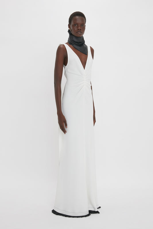 A person wearing a sleek, sleeveless Exclusive V-Neck Gathered Waist Floor-Length Gown In Ivory by Victoria Beckham stands against a plain white background. They also wear a black scarf wrapped around their neck, enhancing the gown’s elegant midi silhouette.