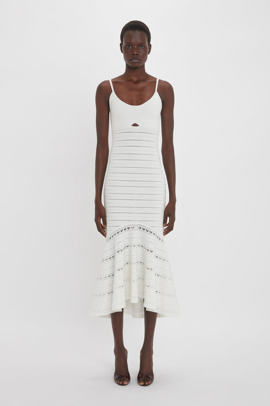 A person stands against a plain white background wearing a white dress with cut-out details, specifically the Cut-Out Detail Cami Dress In White by Victoria Beckham, which is sleeveless and calf-length with a flared hem. The individual pairs it perfectly with black, open-toe shoes.