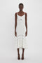 A person stands against a plain white background wearing a white dress with cut-out details, specifically the Cut-Out Detail Cami Dress In White by Victoria Beckham, which is sleeveless and calf-length with a flared hem. The individual pairs it perfectly with black, open-toe shoes.