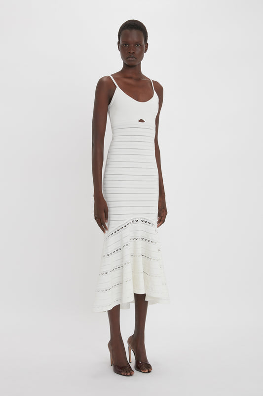 Standing against a plain white background, the person is wearing the Victoria Beckham Cut-Out Detail Cami Dress In White, sleeveless and fitted, with calf-length cutout details at the waist and delicate lace patterns near the hem.