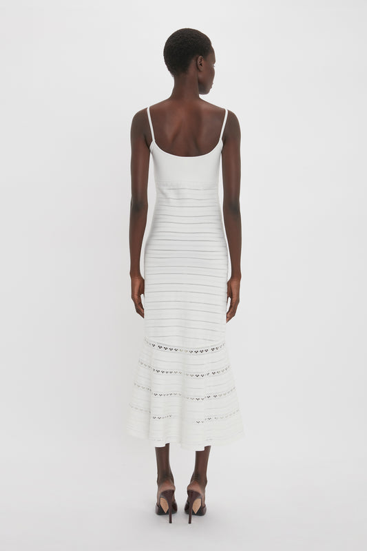 A person is standing with their back facing the camera, wearing a white Cut-Out Detail Cami Dress In White by Victoria Beckham featuring horizontal stripe details and cut-out patterns near the hem, paired with high-heeled shoes.