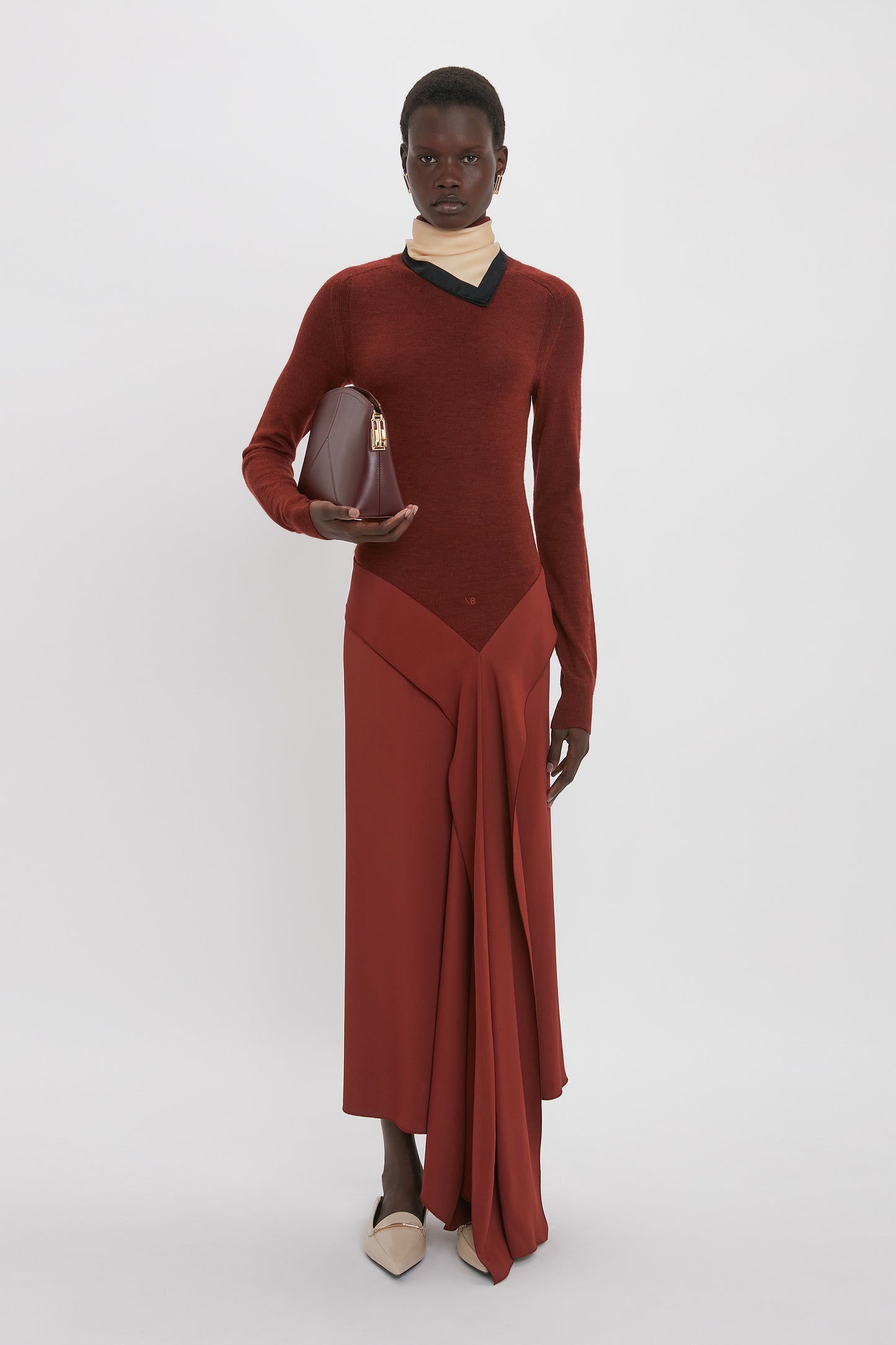 A person stands against a white background wearing the High Neck Tie Detail Dress In Russet by Victoria Beckham, draped in rich red fabric, and holding a burgundy handbag. They also wear beige shoes and a cream-colored neck band.
