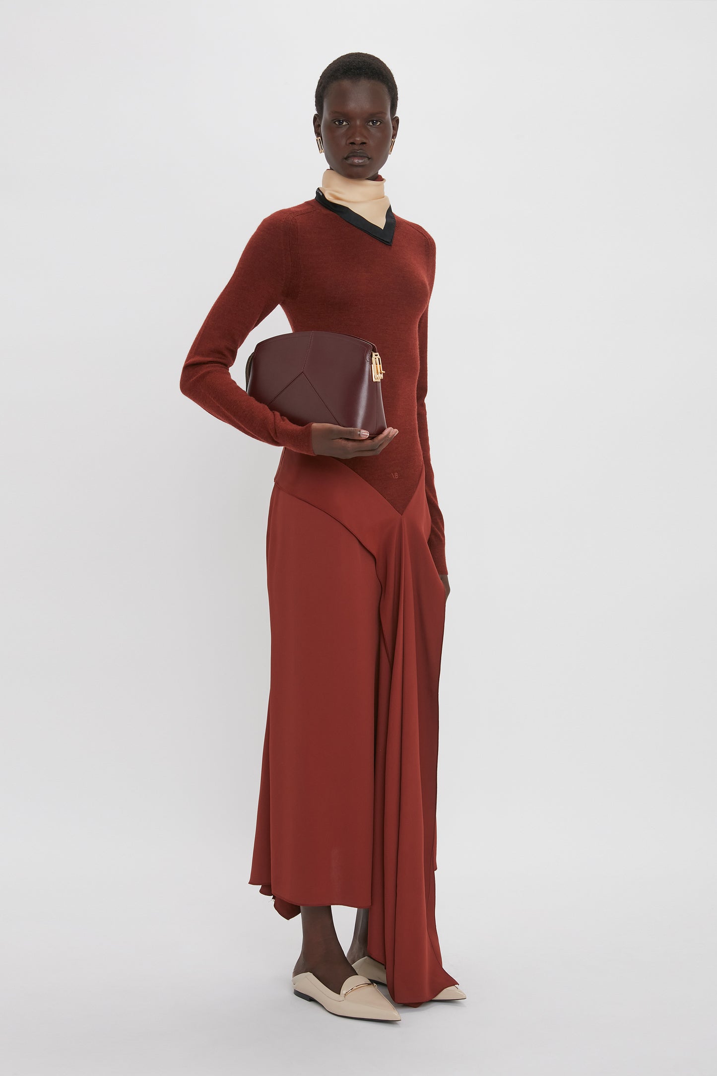 A person stands against a white background wearing the Victoria Beckham High Neck Tie Detail Dress In Russet with a merino wool bodice, long red skirt with an asymmetric hemline, and white shoes, holding a red geometric clutch.