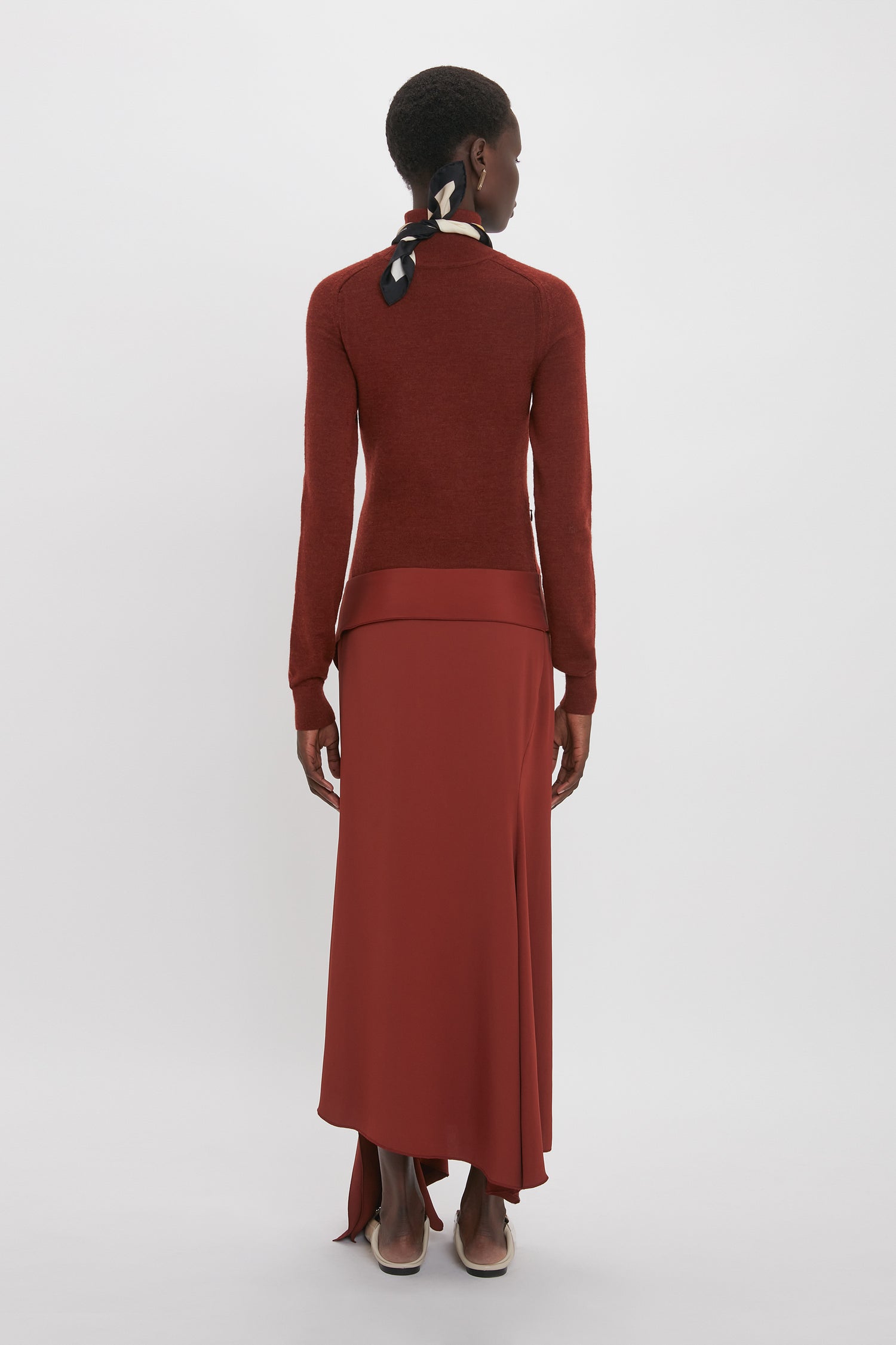 Person with short hair wearing a long-sleeve maroon top and an elegant High Neck Tie Detail Dress In Russet by Victoria Beckham, standing against a plain white background, viewed from the back.
