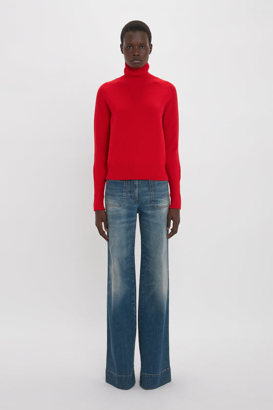 A person stands facing forward wearing a Victoria Beckham Polo Neck Jumper In Red and wide-leg blue jeans against a plain background.