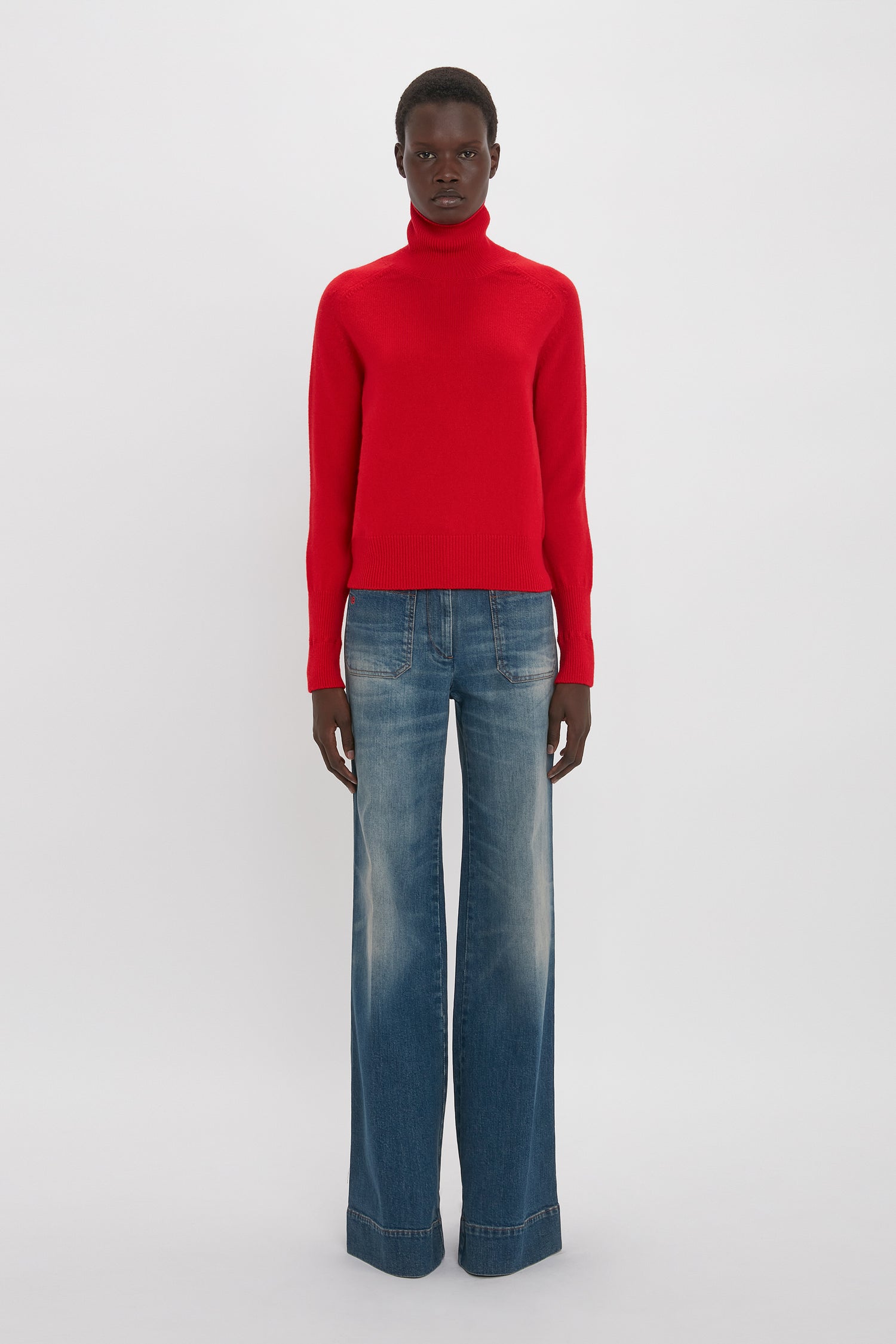 A person standing against a plain background, wearing a red turtleneck sweater and Victoria Beckham's Alina High Waisted Jean In Heavy Vintage Indigo Wash with retro appeal, facing forward.