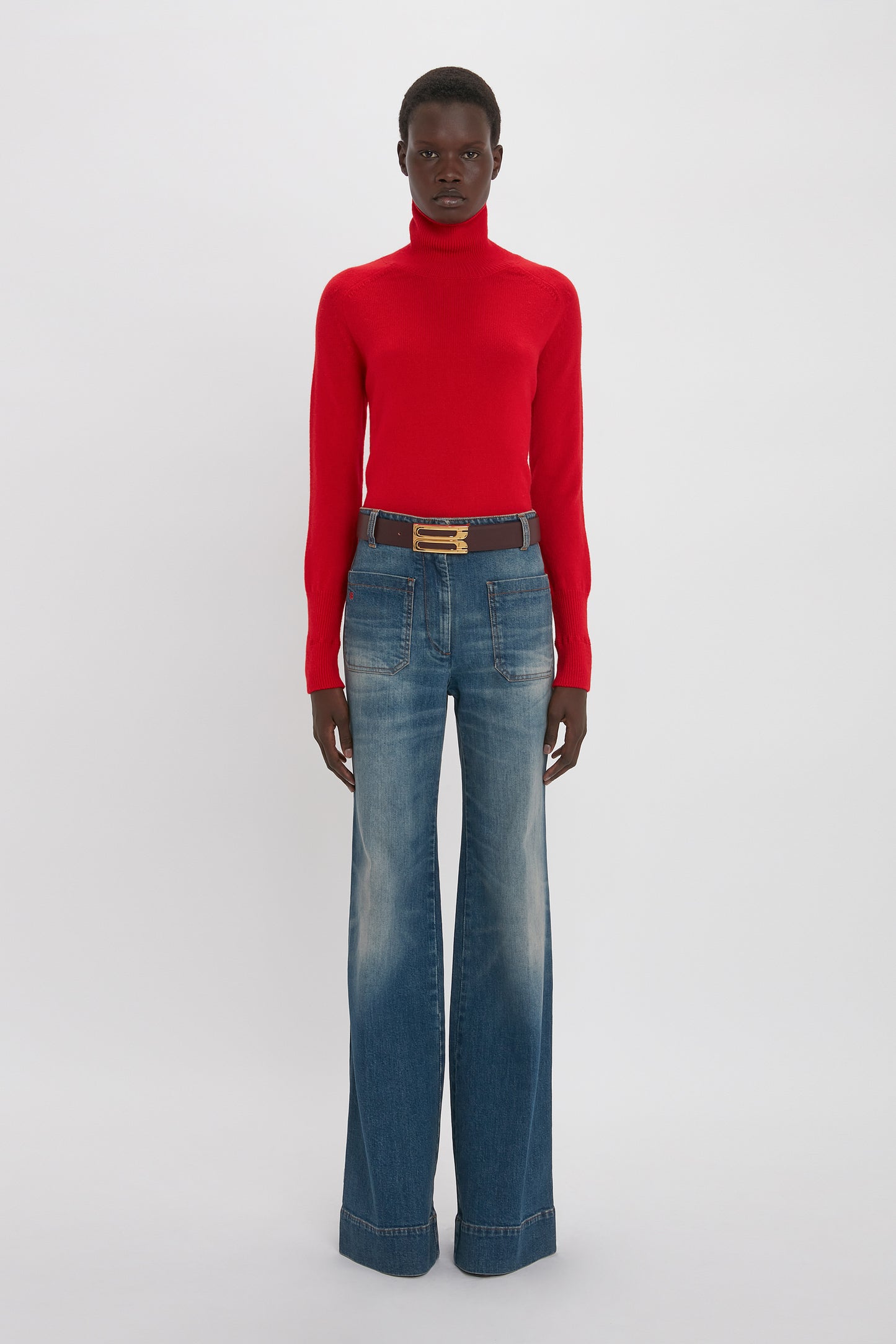 A person stands against a plain background, wearing a luxurious Victoria Beckham Polo Neck Jumper In Red and wide-leg blue jeans with a black belt.