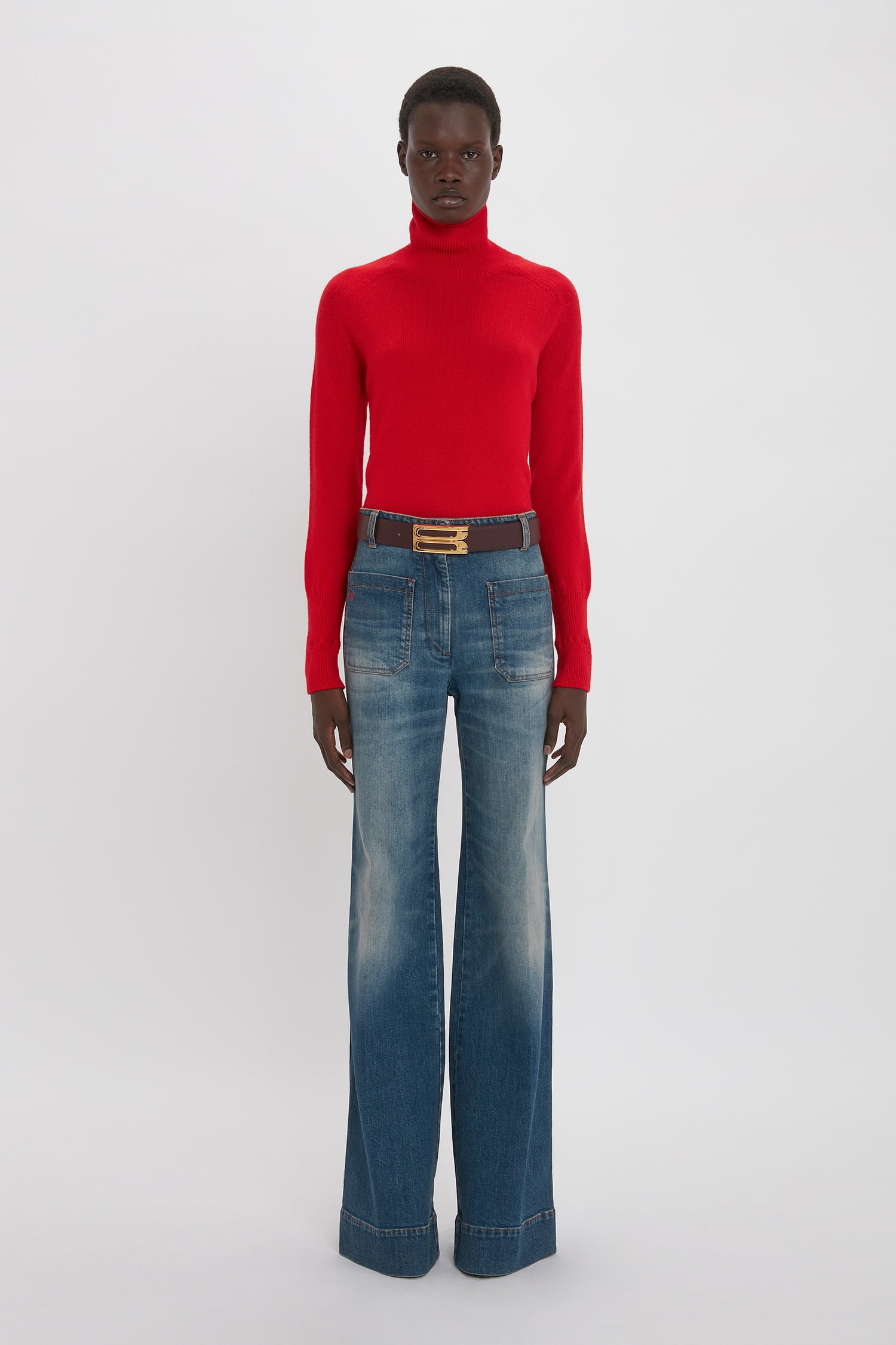 A person stands against a plain background, wearing a luxurious Victoria Beckham Polo Neck Jumper In Red and wide-leg blue jeans with a black belt.