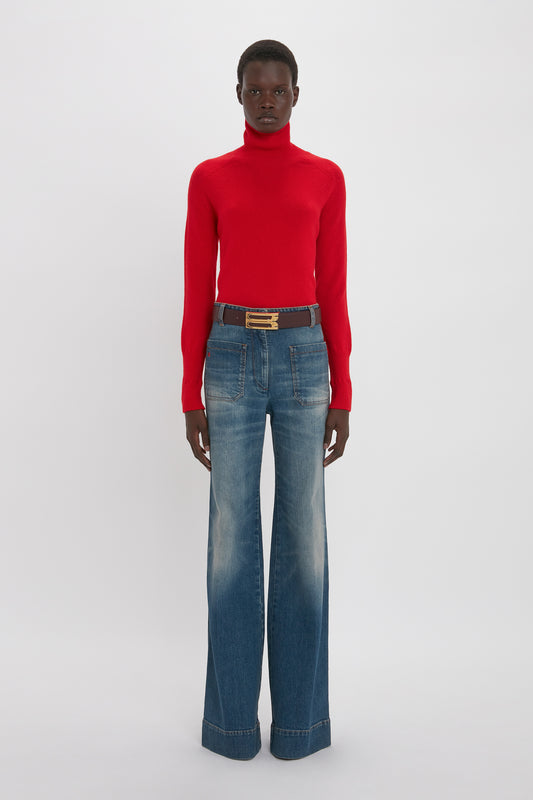 A person stands against a plain background, wearing a luxurious Victoria Beckham Polo Neck Jumper In Red and wide-leg blue jeans with a black belt.