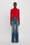 Person standing against a plain background, wearing a red turtleneck sweater and Victoria Beckham Alina High Waisted Jean In Heavy Vintage Indigo Wash exuding retro appeal.