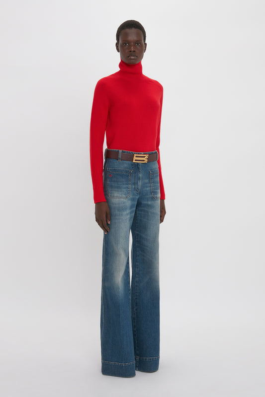 A person stands against a plain background, wearing a Polo Neck Jumper In Red by Victoria Beckham and wide-leg blue jeans with a black belt.