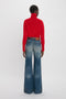 A person wearing a Victoria Beckham Polo Neck Jumper In Red and high-waisted, wide-leg blue jeans stands facing away from the camera against a plain white background.