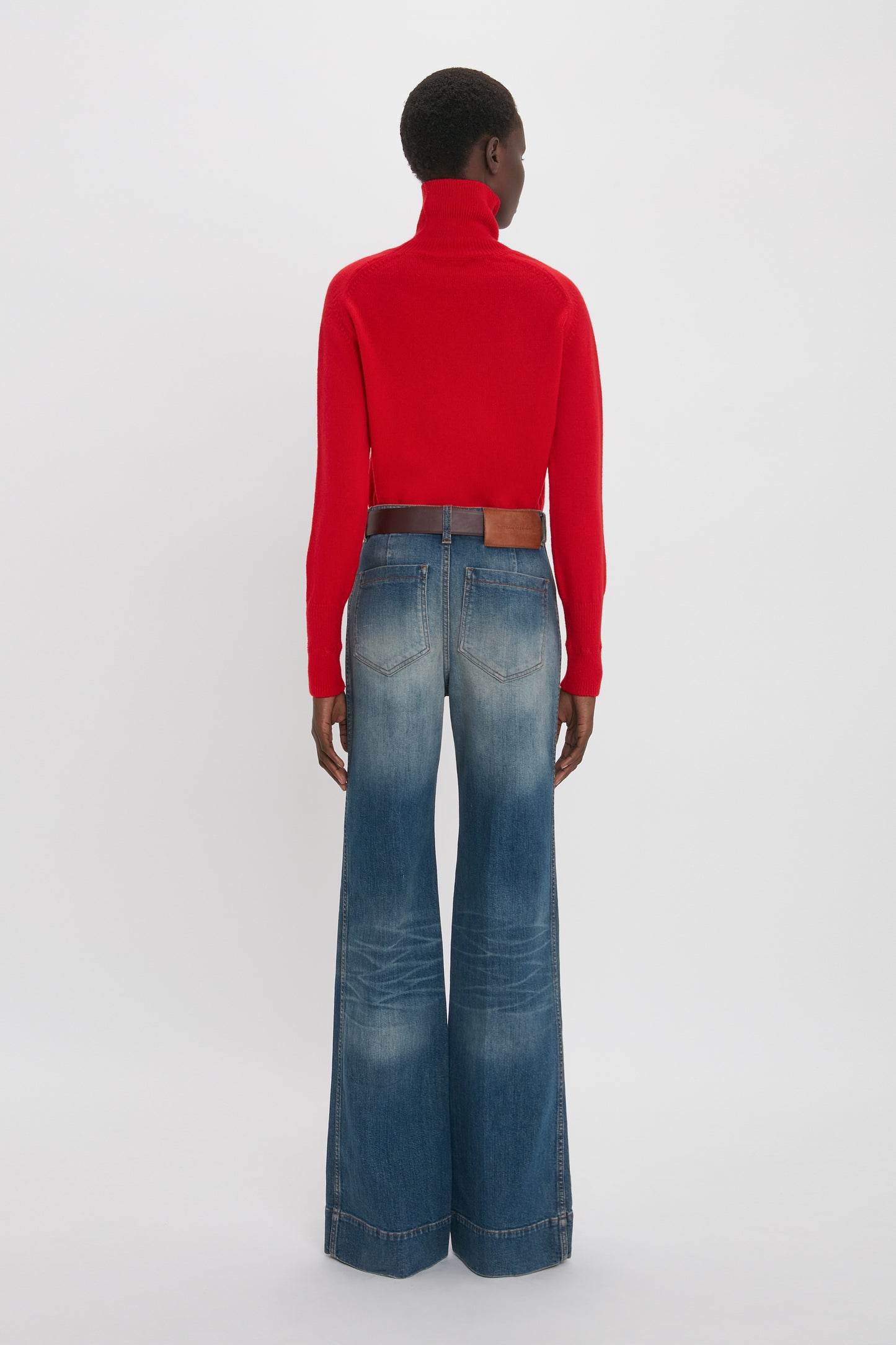 A person stands facing away, exuding retro appeal in a red turtleneck sweater and high-waisted, tailored Alina High Waisted Jean In Heavy Vintage Indigo Wash by Victoria Beckham.