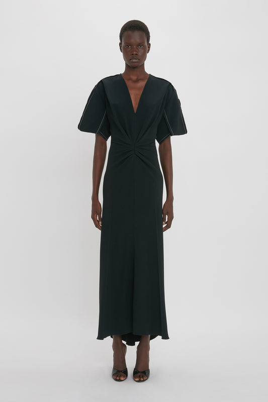 A person stands against a plain white background, wearing a Gathered V-Neck Midi Dress in Black by Victoria Beckham in figure-flattering stretch fabric with short sleeves, paired with black high-heeled sandals, exuding a contemporary edge.