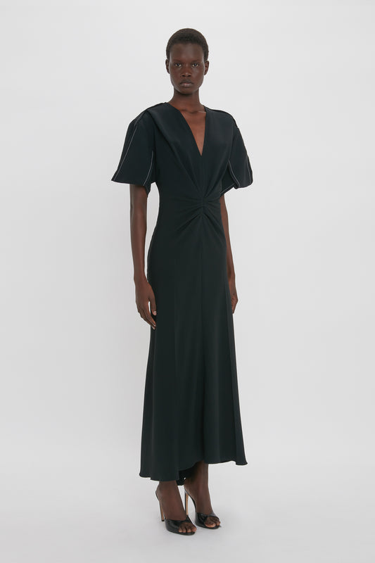 A person stands confidently in a black, short-sleeved, Gathered V-Neck Midi Dress in Black from Victoria Beckham. The floor-length attire features figure-flattering stretch fabric and black high-heeled shoes, exuding a contemporary edge.