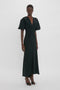 A person stands confidently in a black, short-sleeved, Gathered V-Neck Midi Dress in Black from Victoria Beckham. The floor-length attire features figure-flattering stretch fabric and black high-heeled shoes, exuding a contemporary edge.