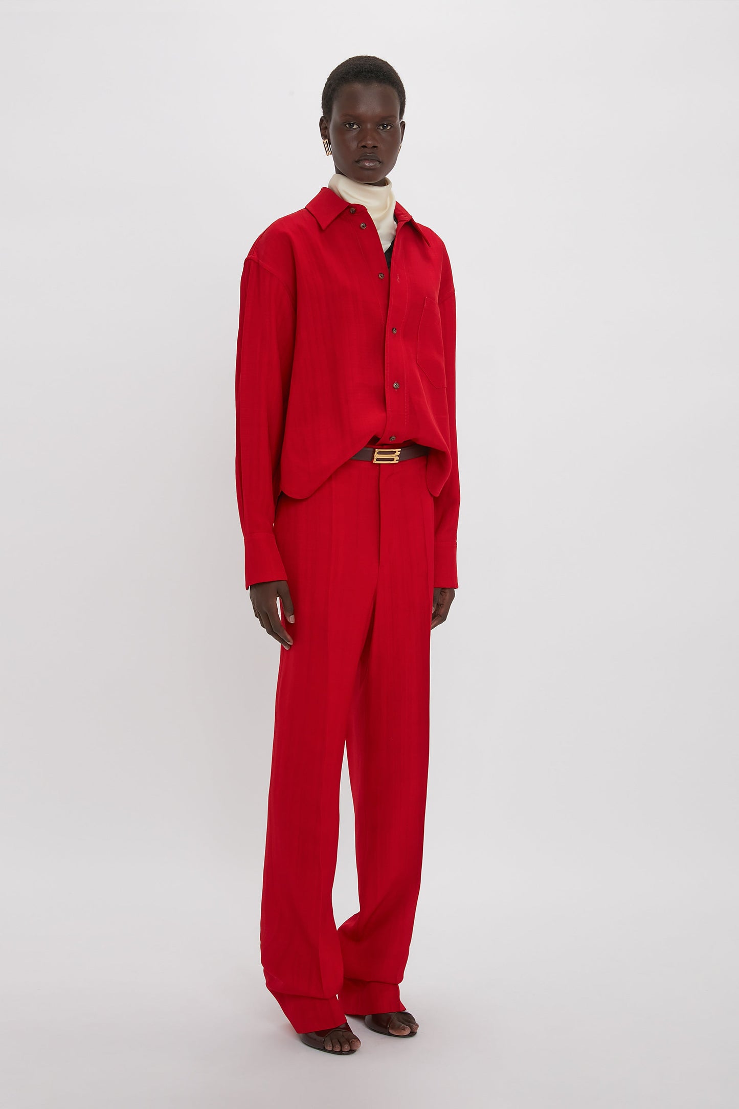 Person standing against a plain background, wearing a bright carmine red long-sleeve shirt and matching Victoria Beckham Tapered Leg Trouser In Carmine, paired with a white inner layer and red sandals, showcasing elements of traditional tailoring.