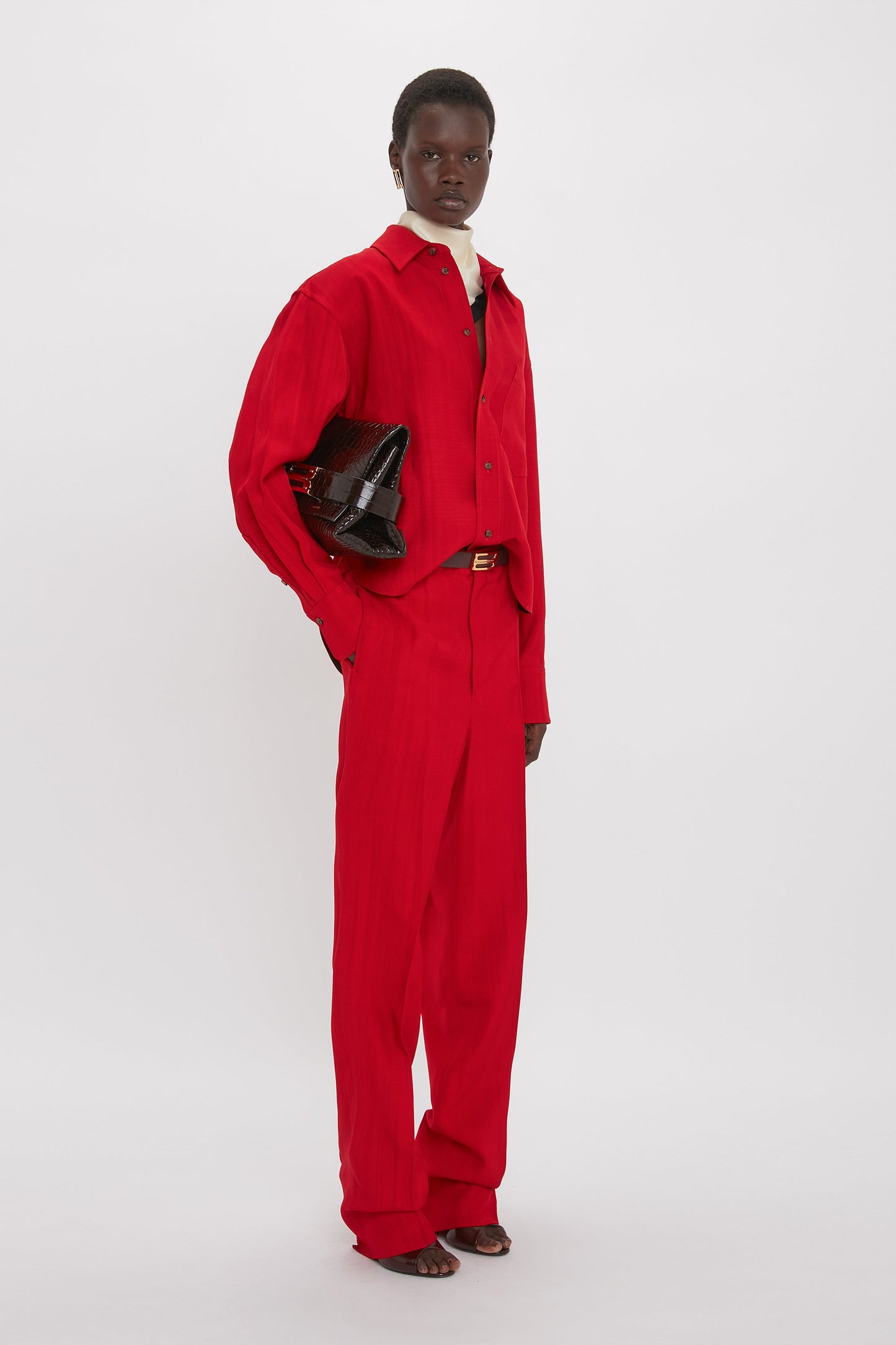 Person wearing a Carmine red button-up shirt and matching Tapered Leg Trouser In Carmine by Victoria Beckham, holding a black clutch bag, standing against a plain white background.