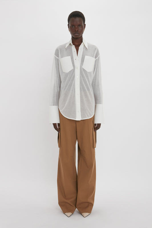 A person stands against a plain white background wearing a relaxed fit, white button-up Pocket Detail Shirt In White by Victoria Beckham and wide-legged brown pants with front patch pockets. They have a neutral expression and their hands are by their sides.
