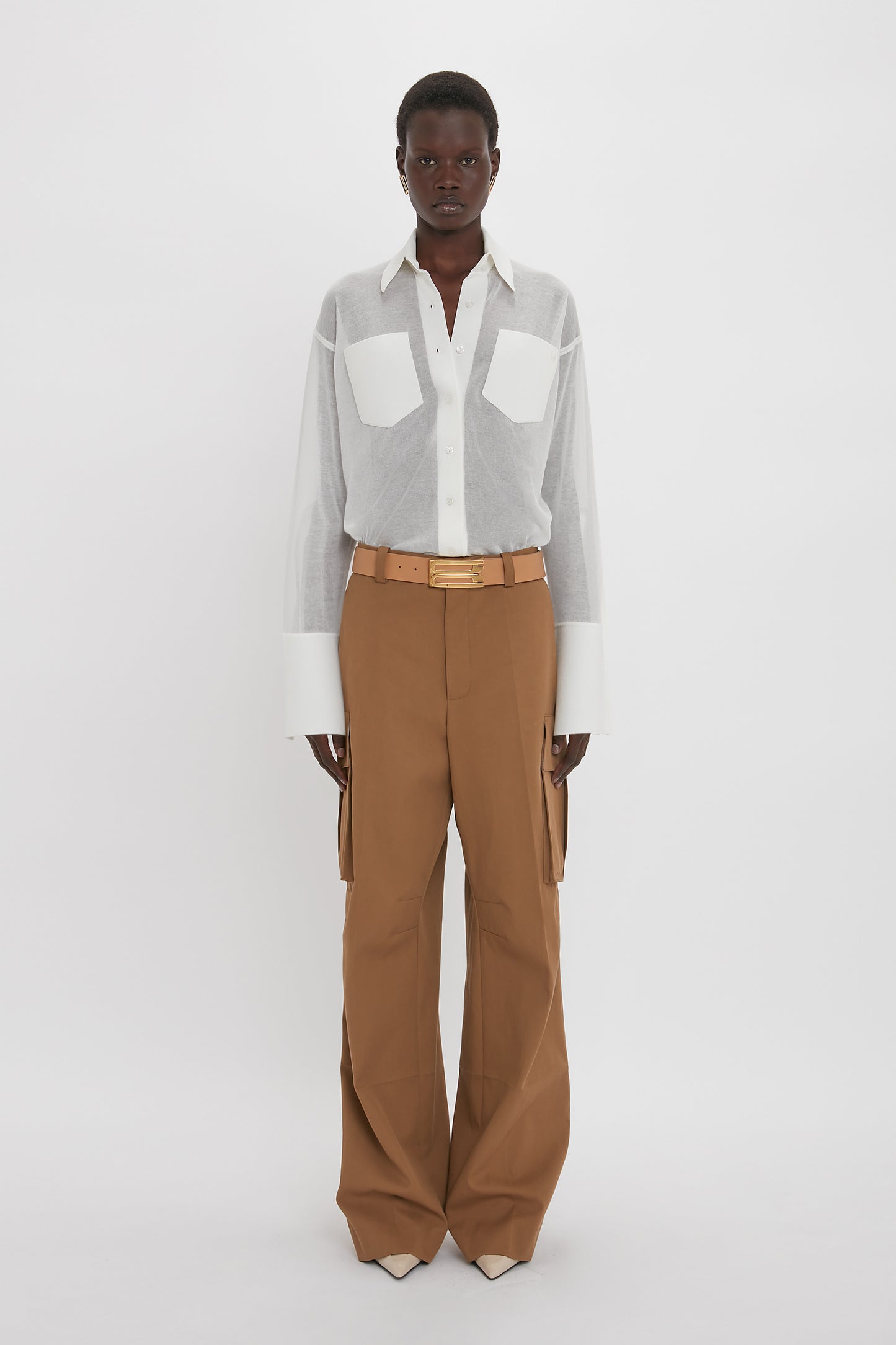 Person standing wearing a white Pocket Detail Shirt In White by Victoria Beckham with front patch pockets and brown high-waisted, wide-leg pants in a relaxed fit. The background is plain white.