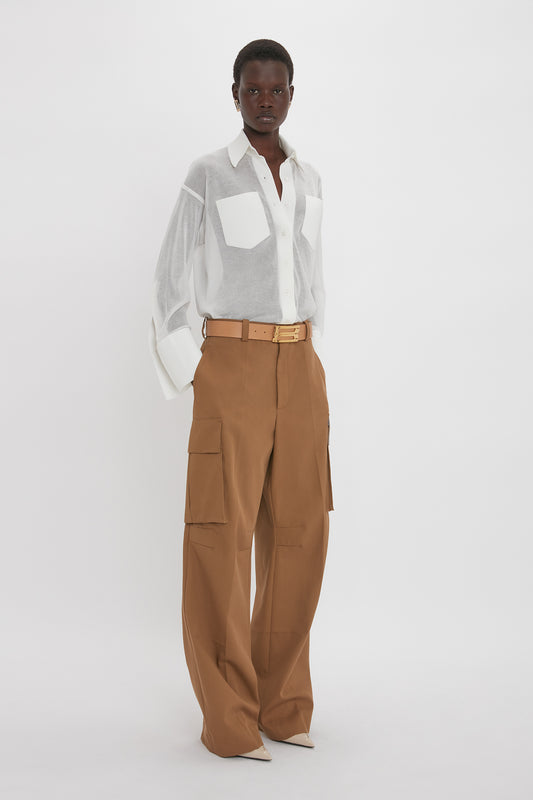 Person standing against a plain white background, wearing a Victoria Beckham Pocket Detail Shirt In White with front patch pockets and brown belted, wide-leg cargo pants with heels.