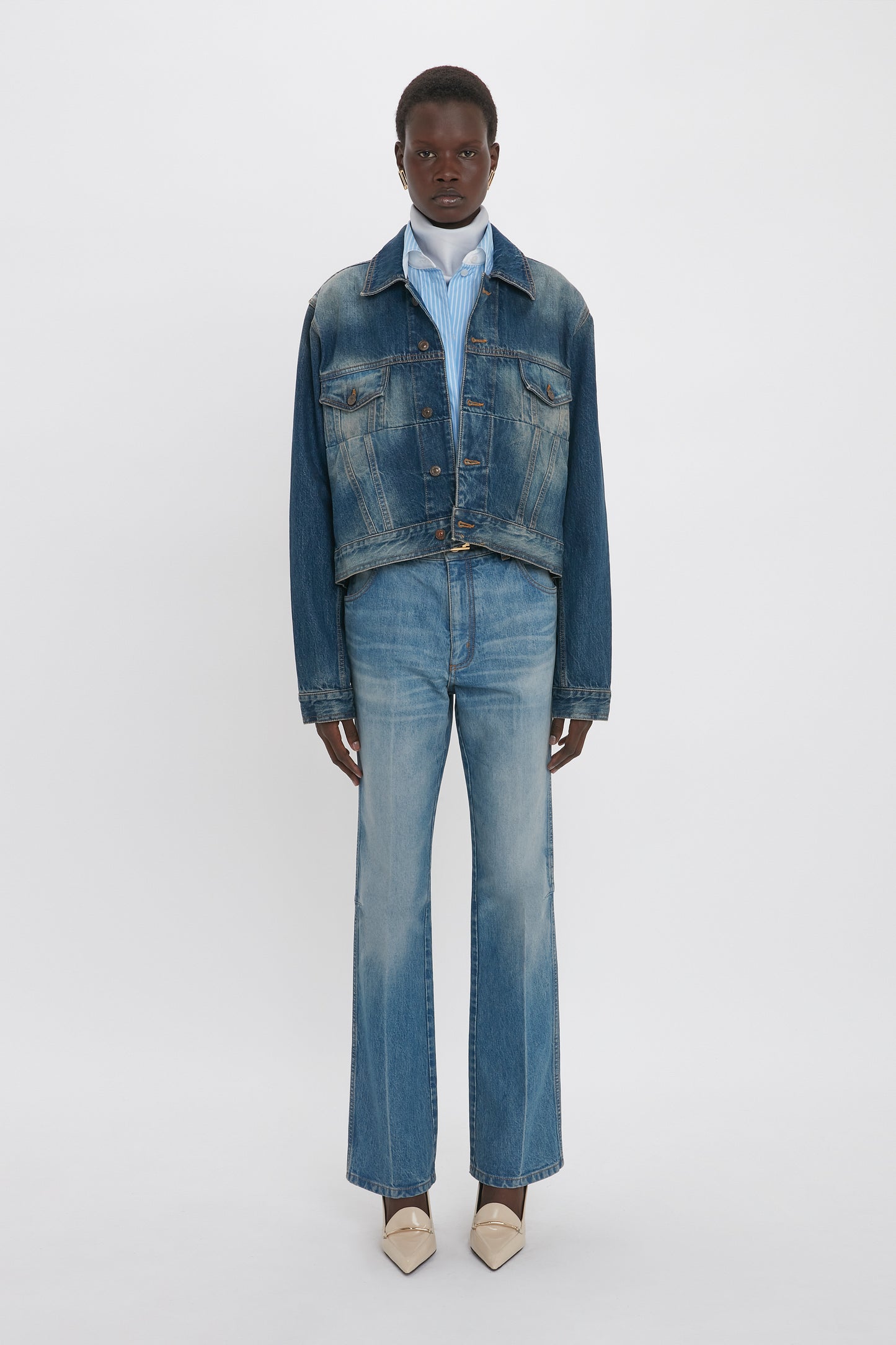 Person wearing a luxury Cropped Denim Jacket In Heavy Vintage Indigo Wash, blue and white striped shirt, blue jeans, and beige pointed shoes stands against a plain white background, embodying the chic style of Victoria Beckham.