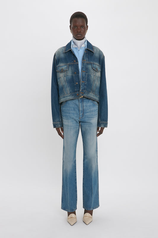 Person wearing a luxury Cropped Denim Jacket In Heavy Vintage Indigo Wash, blue and white striped shirt, blue jeans, and beige pointed shoes stands against a plain white background, embodying the chic style of Victoria Beckham.