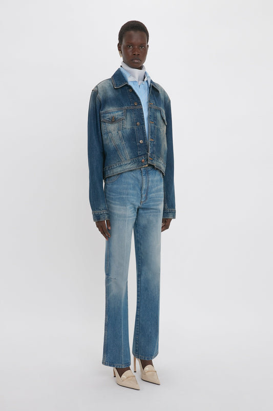 A person stands in front of a plain background wearing a denim jacket, Relaxed Flared Jean In Broken Vintage Wash by Victoria Beckham with deconstructed detailing, a light blue collared shirt, and beige pointed shoes.