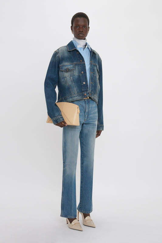 A person stands wearing a Victoria Beckham Cropped Denim Jacket In Heavy Vintage Indigo Wash and jeans, holding a beige clutch. The background is plain white, emphasizing the sleek ensemble reminiscent of Victoria Beckham's signature style.