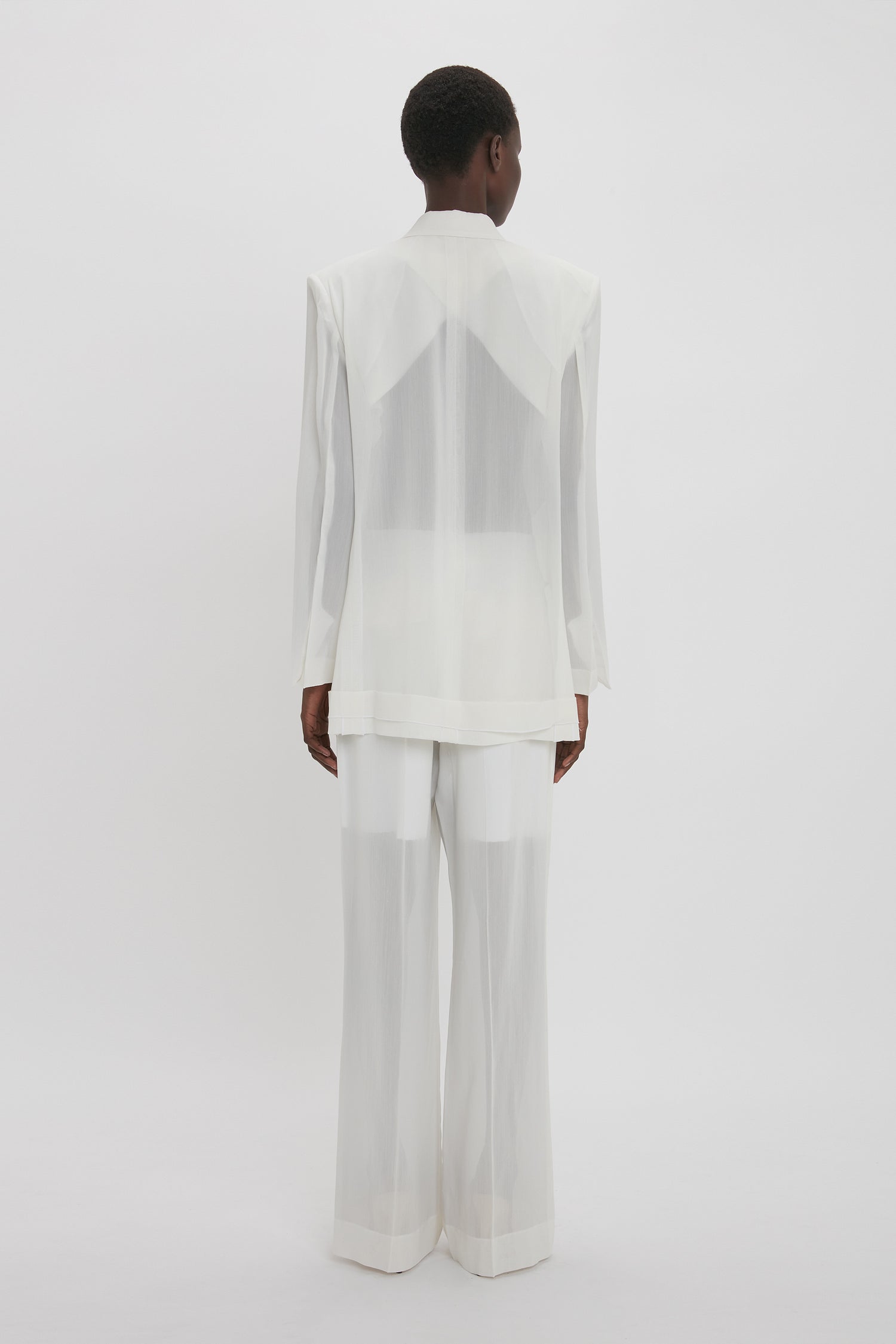 A person with short hair is standing facing away, wearing a white, semi-transparent Fold Detail Tailored Jacket In White by Victoria Beckham and matching white trousers with modern flair against a plain white background.