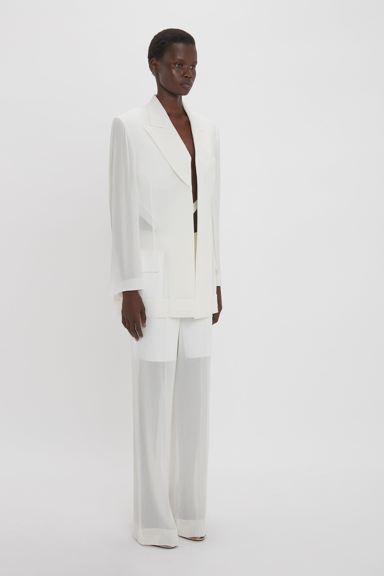 Person wearing a white suit with a long jacket and wide-leg trousers, made from featherweight wool, stands against a plain white background, exuding modern flair in the Fold Detail Tailored Jacket In White by Victoria Beckham.